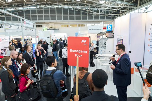Guided trade fair tour with a group of visitors and a guide speaking into a microphone.