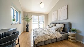 Helles Schlafzimmer mit einem grauen Bett, neutraler Bettwäsche, moderner Einrichtung, Holzböden, einem Fenster, einem Schreibtisch und einer Glastür, durch die Tageslicht hereinströmt.