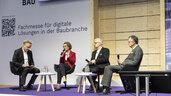 Four people are sitting in a panel discussion on the stage at digitalBAU. A woman in a red blazer is speaking into the microphone.