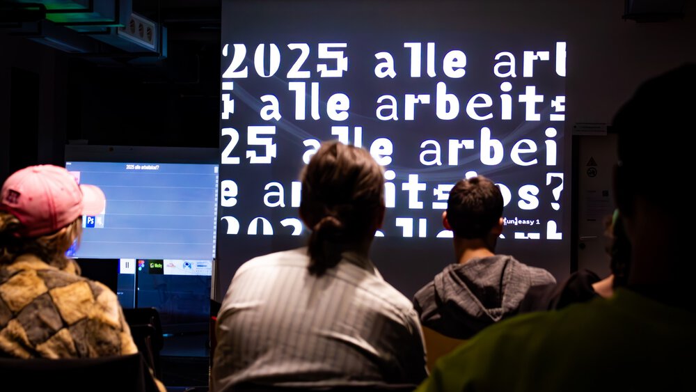 People watch a presentation in which the text “2025 all work” is projected onto a screen in bold print.