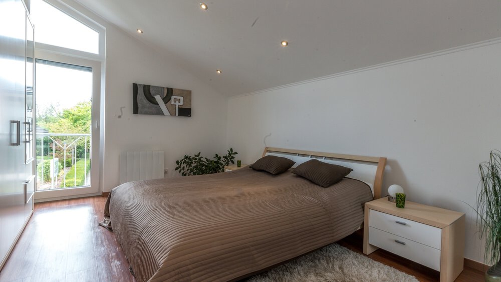 Helles Schlafzimmer mit großem Bett, brauner Bettwäsche, weißen Wänden und einem kleinen Nachttisch neben einem Fenster mit Blick auf den Garten.