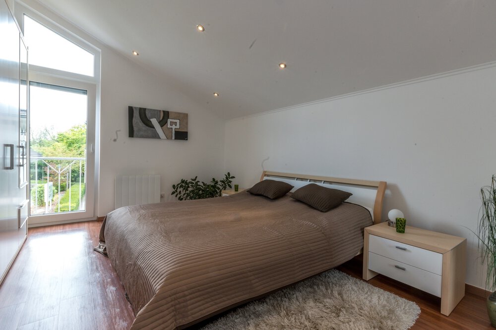 Helles Schlafzimmer mit großem Bett, brauner Bettwäsche, weißen Wänden und einem kleinen Nachttisch neben einem Fenster mit Blick auf den Garten.