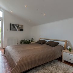 Helles Schlafzimmer mit großem Bett, brauner Bettwäsche, weißen Wänden und einem kleinen Nachttisch neben einem Fenster mit Blick auf den Garten.