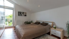 Helles Schlafzimmer mit großem Bett, brauner Bettwäsche, weißen Wänden und einem kleinen Nachttisch neben einem Fenster mit Blick auf den Garten.