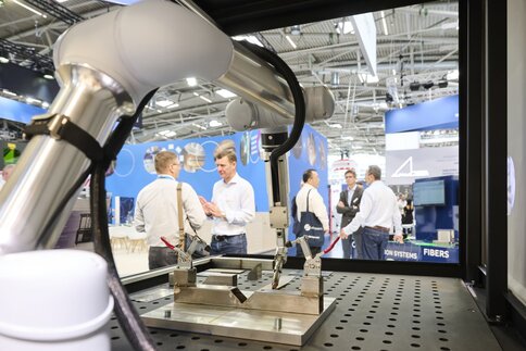 Industrieroboterarm in einer Messehalle, der an einer Metallvorrichtung arbeitet, um den Einsatz moderner Lasersysteme in der Fertigungstechnik zu demonstrieren.