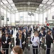 A crowd of people streaming through the ticket barriers in the large entrance hall of the trade fair.