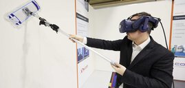 Ein Mann im Anzug trägt ein Headset und wischt mit einem Wischmopp über die Wand eines Standes auf der productronica.