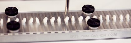 Several white, round pills lie next to each other, each in a matching hollow. One pill is sucked in by a round metal tube.