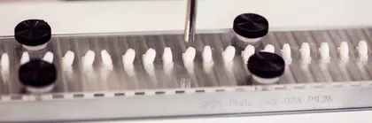 Einige weiße, runde Pillen liegen nebeneinander, jeweils in einer passenden Mulde. Eine Pille wird von einem runden Metallröhrchen angesaugt.