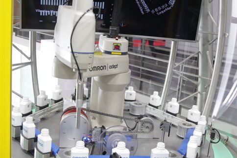 Roboterarm um den sich kleine weiße Flaschen auf einem hexagonalen Fließband bewegen. Im Hintergrund ist ein Monitoren mit Testwerten.