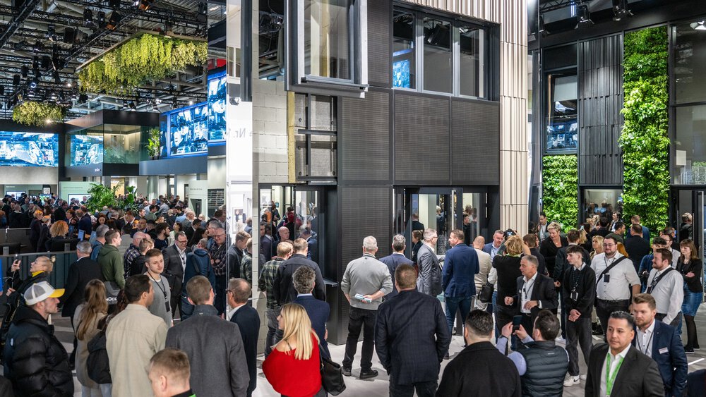 A large crowd gathers in an indoor exhibition with modern architecture and green walls.