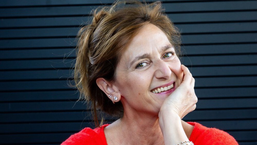 A smiling woman with dark blonde hair tied up is wearing a loose-fitting red top, a decorative bracelet and earrings.