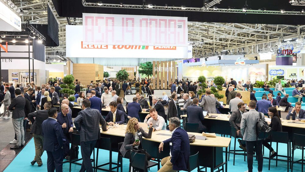 Area with tables and chairs where people socialize, in a large exhibition hall with numerous exhibition stands in the background.