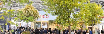 Viele Menschen an einem Platz in einer großen Messehalle, mit vielen Bäumen, Büschen und Sitzgelegenheiten aus Holz.