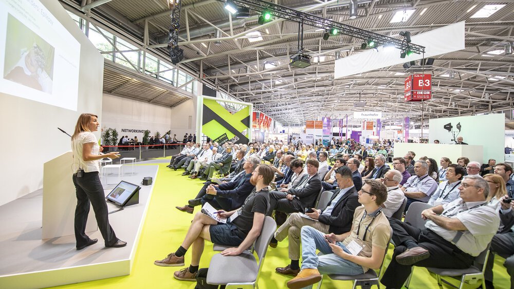 Bei einer großen, modernen Konferenz in einem geschlossenen Raum hält eine Frau vor einem sitzenden Publikum einen Vortrag.