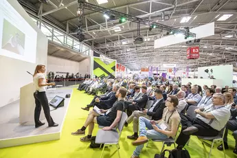 Bei einer großen, modernen Konferenz in einem geschlossenen Raum hält eine Frau vor einem sitzenden Publikum einen Vortrag.