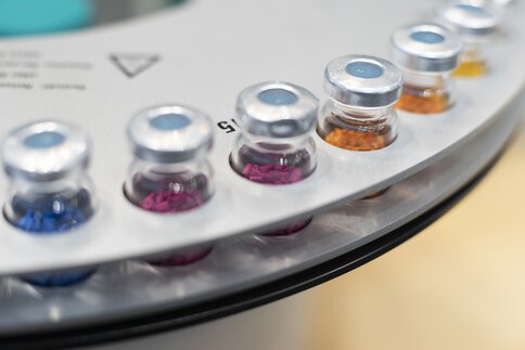 Nahaufnahme eines Analysegeräts: In runden Halterungen sind im Kreis mehrere Fläschchen mit Aluminium-Septumverschluss (Vials) mit bunten Körnern gefüllt angeordnet.