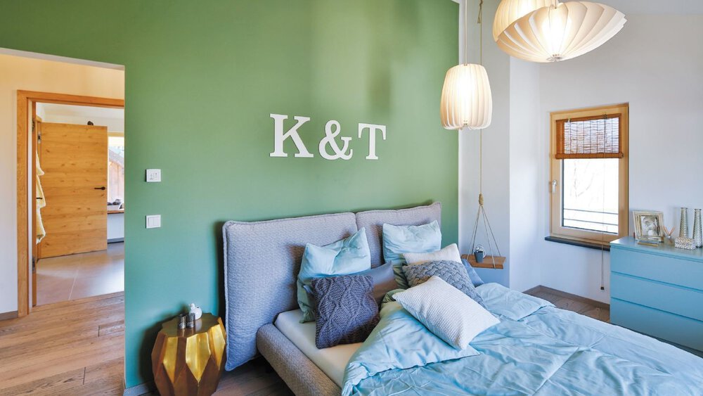 Ein gemütliches Schlafzimmer mit einer grünen Akzentwand, einem grauen Bett und blaugrünen Kissen. An der Wand über dem Bett stehen die Initialen 