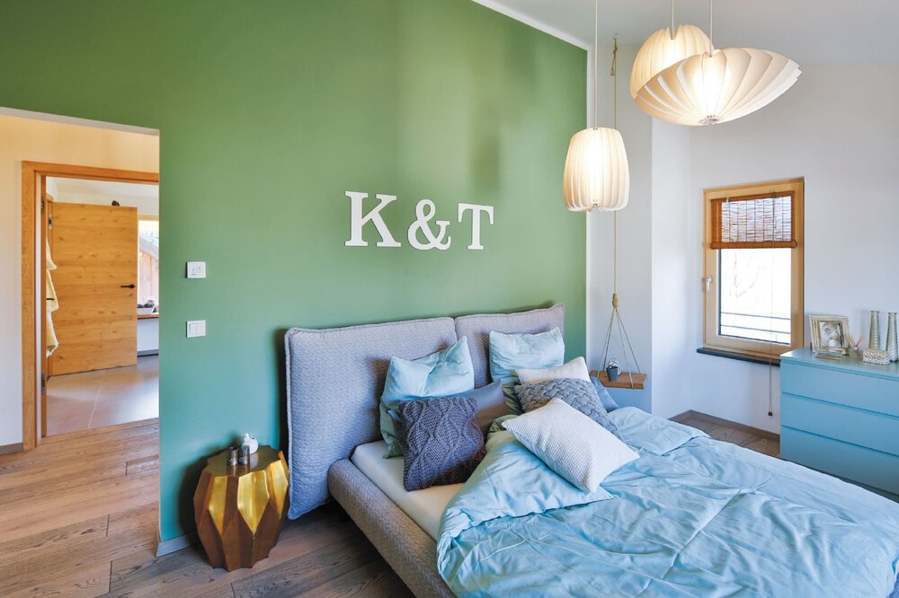 Ein gemütliches Schlafzimmer mit einer grünen Akzentwand, einem grauen Bett und blaugrünen Kissen. An der Wand über dem Bett stehen die Initialen 