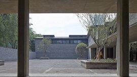 Betonsäulen rahmen einen Innenhof mit Ziegelpflaster, Bäumen und modernen grauen Backsteingebäuden ein.