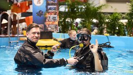 Three people in diving suits stand in a pool of water at the f.re.e exhibition.
