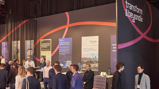 Besucherinnen und Besucher unterhalten sich im Bereich „transform & beyond by EXPO REAL“. Mehrere Informationsstände mit farbigen Bannern präsentieren Themen rund um Energieeffizienz, Dekarbonisierung und nachhaltige Immobilienlösungen. Die Atmosphäre ist geschäftig, mit vielen Gesprächen zwischen Fachleuten aus der Immobilienbranche.