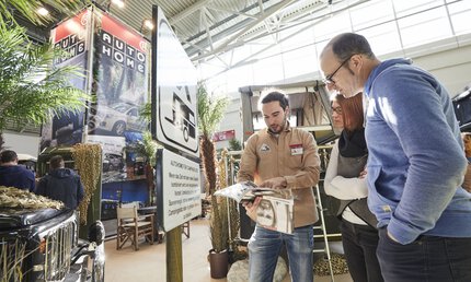 Nahaufnahme, ein Mann steht in einer Messehalle neben einem Camping-Straßenschild und zeigt etwas in einem Katalog