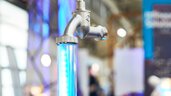 Nahaufnahme eines Wasserhahns der auf einer beleuchteten Plexiglassäule montiert ist, die den Wasserstrahl symbolisieren soll. Im Hintergrund sind unscharf weitere Messestände zu sehen.