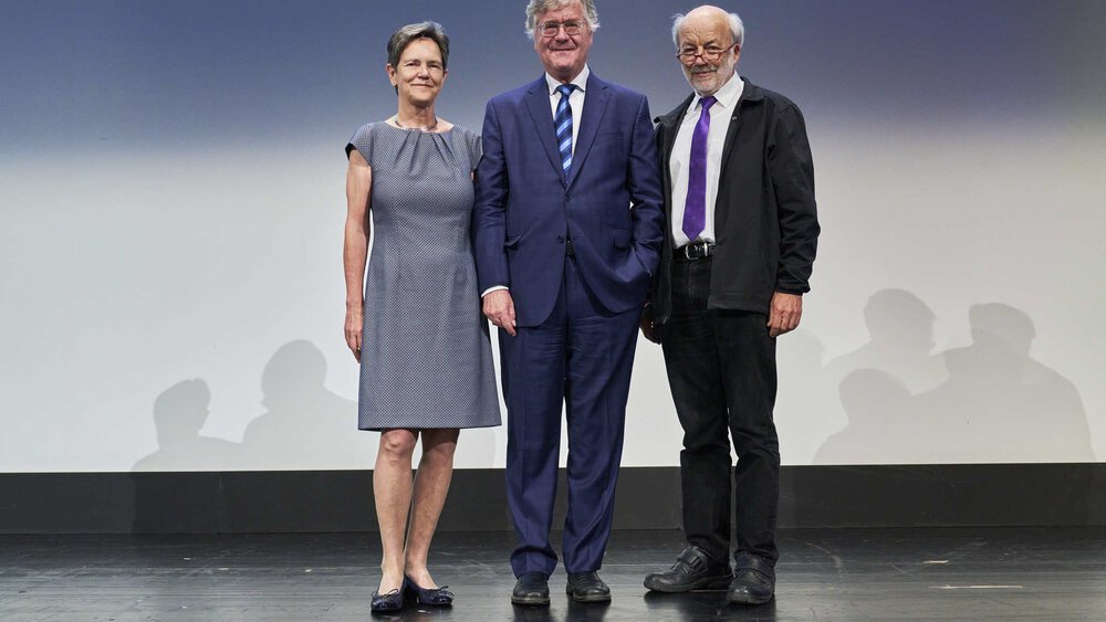 Three people are standing on a stage with a neutral background: a woman in a gray dress and two men in suits.
