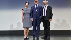 Three people are standing on a stage with a neutral background: a woman in a gray dress and two men in suits.