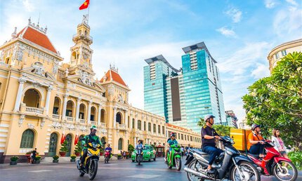 People ride motorcycles past a historic yellow building and modern skyscrapers in a city in Vietnam.
