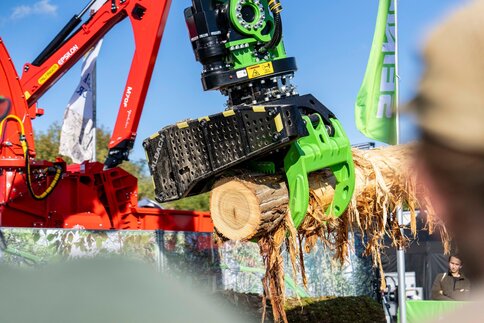 Green grapple of a forestry machine holding a tree trunk during an outdoor demonstration.