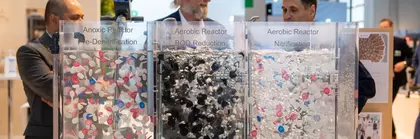 Drei Männer diskutieren an einem Ausstellungsstand hinter drei transparenten Reaktionsmodellen zur Abwasseraufbereitung.