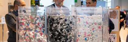 Three men discuss wastewater treatment at an exhibition stand behind three transparent reaction models.