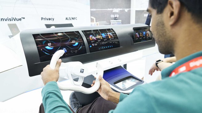 A man sits at a steering wheel in front of a futuristic dashboard with several digital displays.