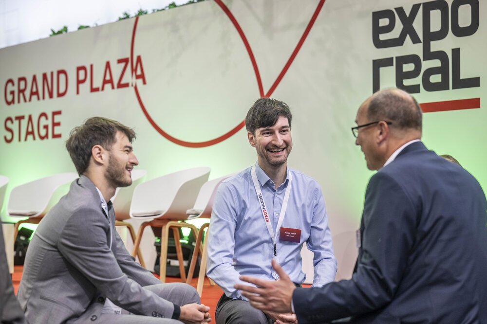 Three visitors are talking in front of the Grand Plaza Stage at EXPO REAL, exchanging ideas in a relaxed atmosphere.