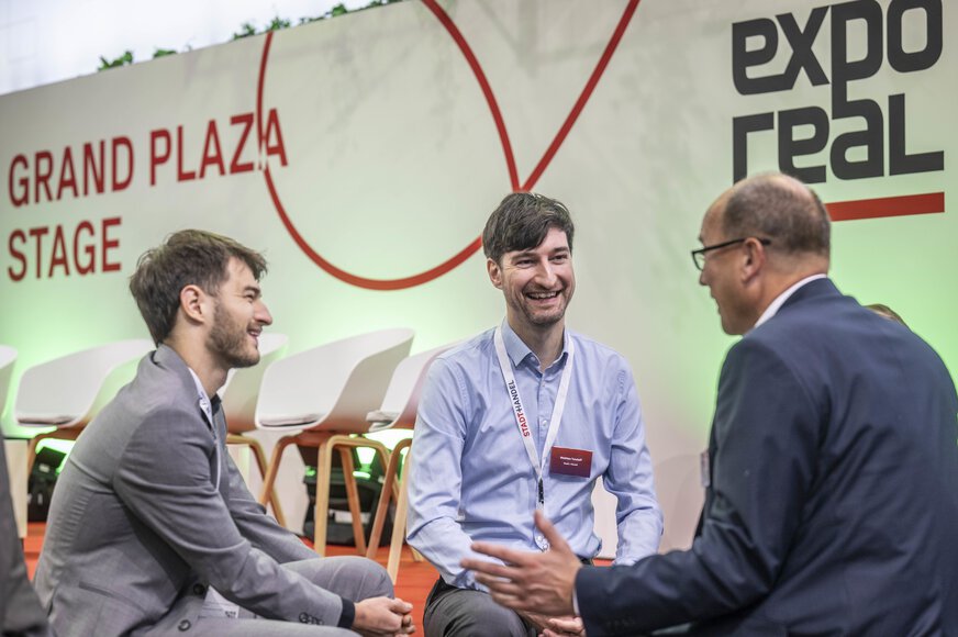 Three visitors are talking in front of the Grand Plaza Stage at EXPO REAL, exchanging ideas in a relaxed atmosphere.