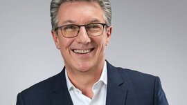 Ein lächelnder Mann mit kurzen grauen Haaren trägt  eine schwarze Brille und ein weißes Oberhemd mit einem blauen karierten Jackett darüber.