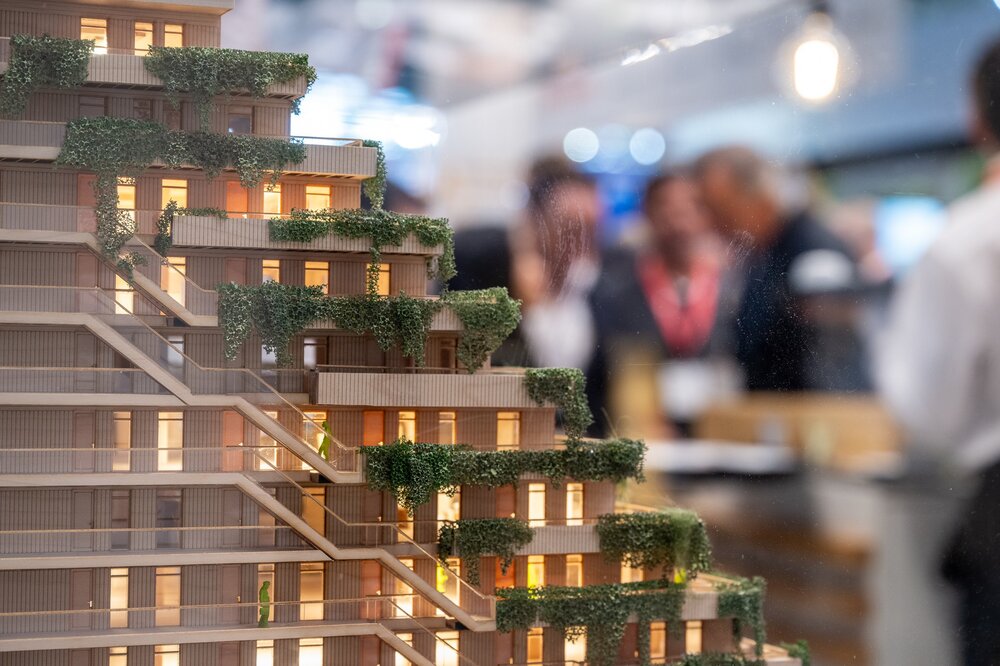 Ein beleuchtetes Architekturmodell eines stufenförmigen, mehrstöckigen Gebäudes mit Dachbegrünung und verschwommenen Menschen im Hintergrund.