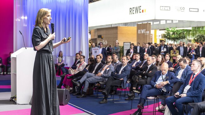 Eine Frau hält auf der EXPO REAL in München einen Vortrag vor einem großen Publikum. Sie steht mit Mikrofon und Präsentationsfernbedienung auf der Bühne und spricht konzentriert. Das Publikum, bestehend aus Fachbesuchern in Businesskleidung, hört aufmerksam zu. Im Hintergrund sind Messestände und ein großes REWE-Logo sichtbar.