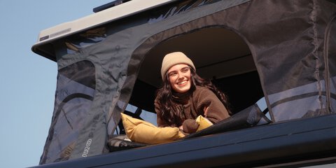 Eine Frau schaut lächelnd aus einem Dachzelt und genießt das Outdoor-Erlebnis. Das Bild steht für modernes Camping mit Dachzelt und passt zur Podcast-Folge über Dachzelte auf der f.re.e.