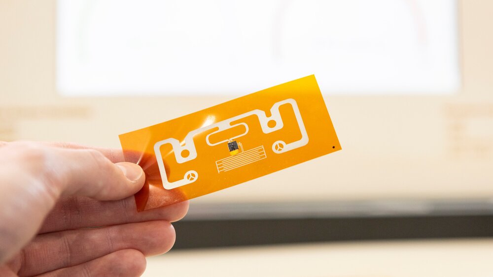 Eine Hand hält eine simple kleine orangen-weiße Leiterplatine zwischen den Fingern. Der Hintergrund ist unscharf.