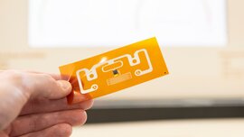 Eine Hand hält eine simple kleine orangen-weiße Leiterplatine zwischen den Fingern. Der Hintergrund ist unscharf.