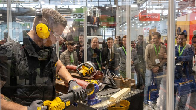 At one stand, a man in protective clothing demonstrates an electric saw while an interested audience watches from behind the protective screens.