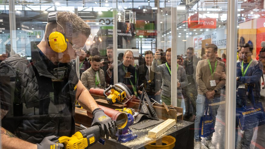 Auf der BAU demonstiert ein Mann in Schutzkleidung eine Elektrosäge, während ihm ein interessiertes Publikum hinter den Schutzscheiben zusieht.