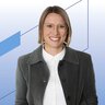 Porträt einer Person mit kurzem, geradem Haar und runder Brille, die frontal in die Kamera blickt. Die Person trägt ein weißes Hemd unter einer dunklen Jacke und steht vor einem hellblauen Hintergrund mit dezenten geometrischen Linien und Formen.