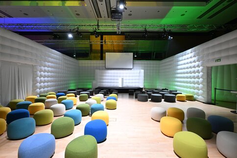 A modern event space with light-colored wood flooring, colorful round stools, white upholstered walls, and a stage with a projection screen at the front.