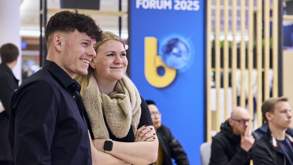 Zwei junge Messebesucher:innen lächeln vor „bauma FORUM 2025“-Hintergrund.