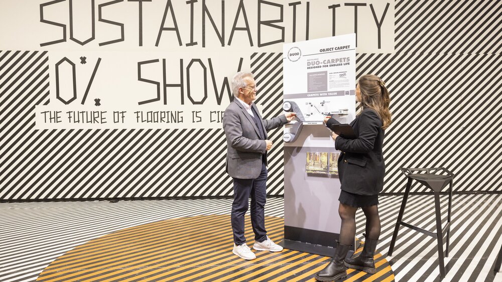A presentation area with a black and white line pattern on the floor and wall. A man and a woman are standing in front of a display board and looking at carpet samples.