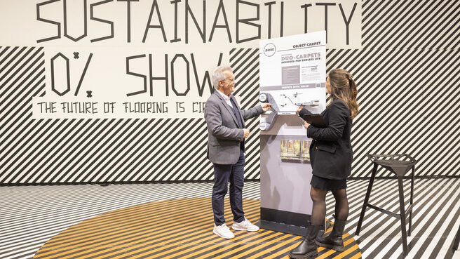 A presentation area with a black and white line pattern on the floor and wall. A man and a woman are standing in front of a display board and looking at carpet samples.